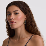 A close-up portrait of a woman with long, wavy brown hair wearing a dark navy blue spaghetti-strap top and a single oval gold earring with a dark stone, set against a plain light gray background.