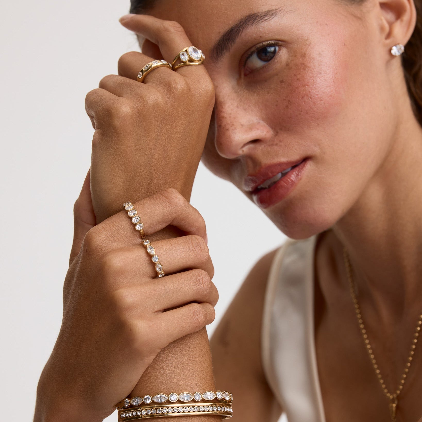 A close-up lifestyle shot of a model wearing multiple gold jewelry pieces, including a ring with a large clear gemstone, several delicate rings, a bracelet, and a necklace, against a clean light gray background.