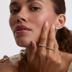 A close-up lifestyle shot of a woman with her hand resting on her cheek, showcasing a delicate silver band ring set with small clear stones and a matching stud earring. The background is a clean, neutral white studio backdrop.