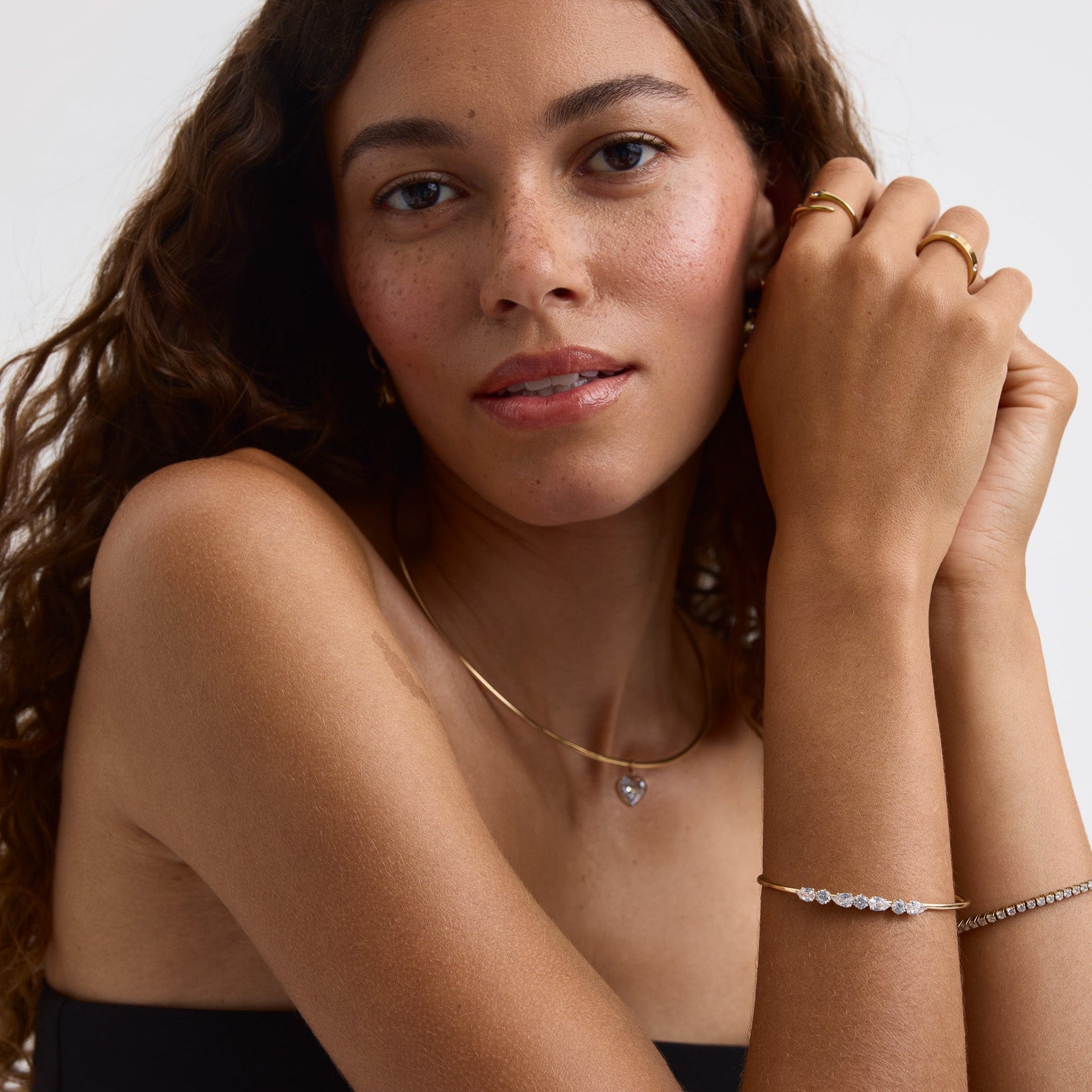 A close-up of a woman with long wavy brown hair wearing delicate gold jewelry, including a thin gold chain necklace with a small heart pendant and gold rings on her fingers, against a plain light background.