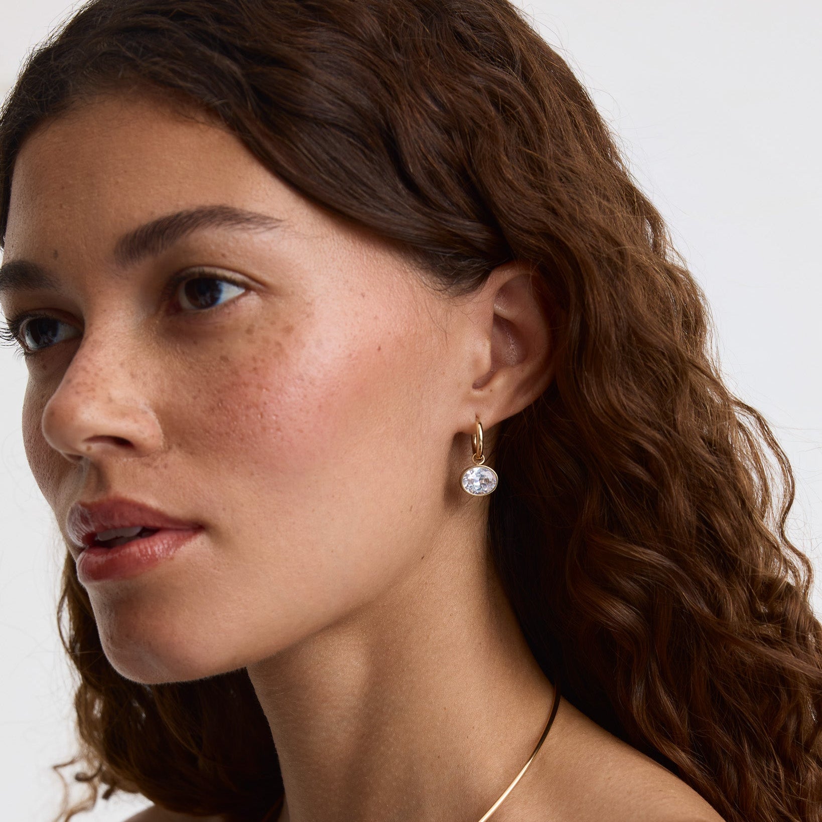 A close-up of a woman with wavy brown hair wearing a delicate gold hoop earring with a clear round gemstone drop, shown against a plain light background.