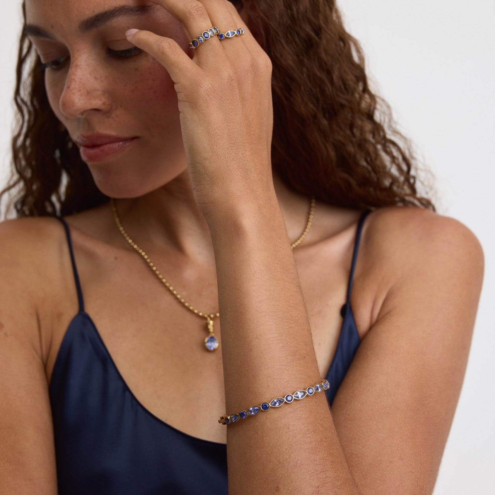 A woman with long wavy brown hair wearing a gold chain necklace with a blue pendant, a gold bracelet, and a silver ring with blue stones, posing against a plain white background.