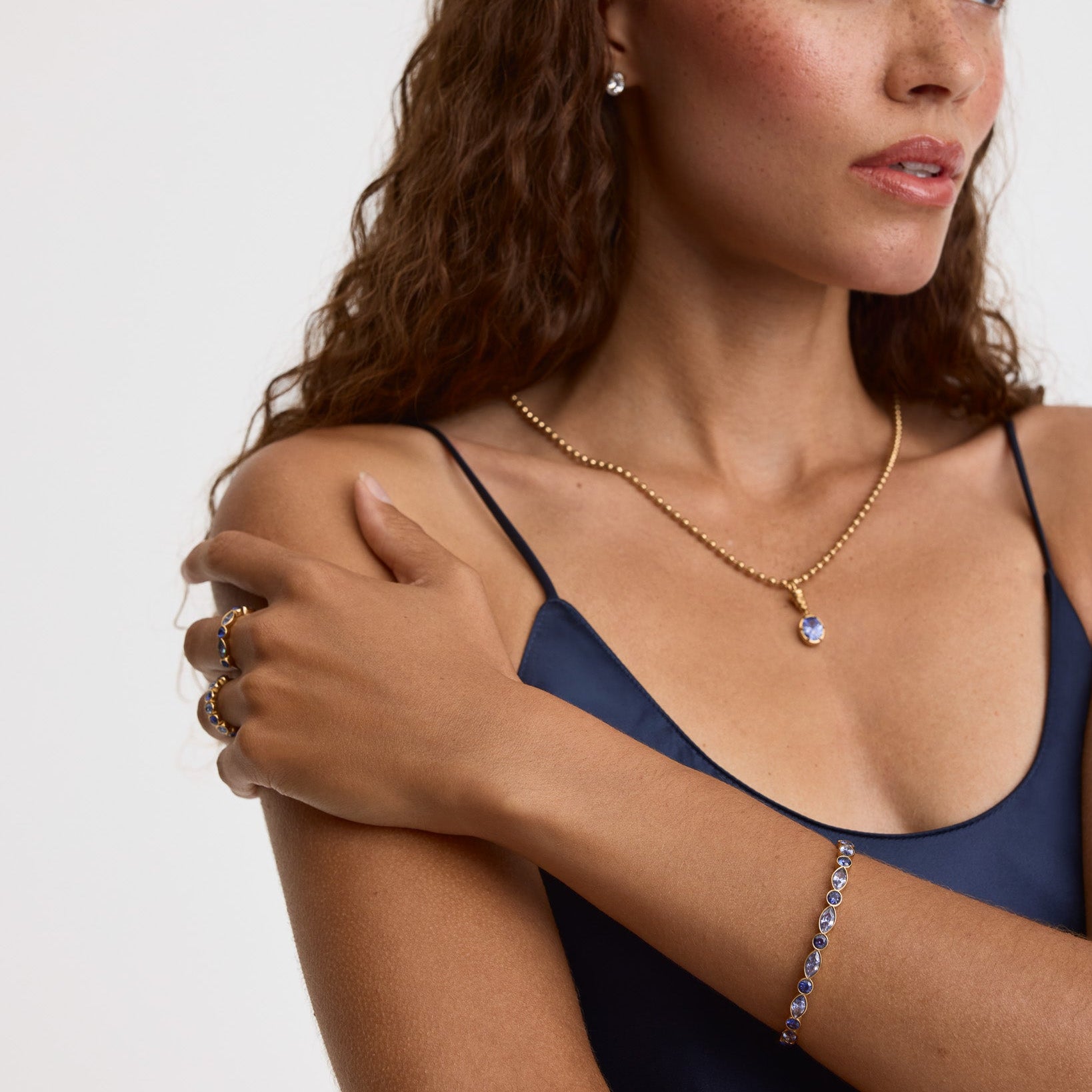 A woman with long wavy brown hair models gold jewelry, including a delicate gold chain necklace with a small light blue teardrop pendant, a matching gold ring, and a gold bracelet with sparkling blue stones, against a clean white background.