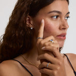 A close-up lifestyle shot of a woman with curly brown hair wearing multiple gold rings with blue gemstones and a gold chain necklace, touching her cheek with her index finger against a light gray background.