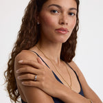 A woman with long, wavy brown hair is shown from the chest up against a plain light background, wearing a delicate gold chain necklace, a gold ring with a clear gemstone, and a gold bracelet.