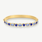 A gold bangle bracelet featuring a row of marquise-cut and round blue gemstones, displayed on a clean white background.