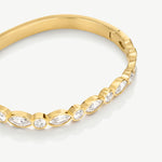 A close-up of a gold bangle bracelet featuring a row of bezel-set marquise and round clear stones, photographed on a clean white background.