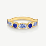 A gold band ring featuring a row of marquise-cut and oval-cut blue gemstones, photographed on a clean, light off-white background.