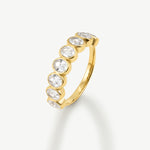 A yellow gold band ring featuring a row of bezel-set round clear gemstones, photographed on a clean off-white background with a soft shadow.