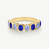 A gold eternity-style ring featuring alternating oval-cut deep blue and light blue gemstones in a bezel setting, shown on a clean off-white background.