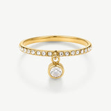 A gold ring featuring a thin band paved with small clear round stones and a dangling bezel-set round clear gemstone, shown on a clean white background.