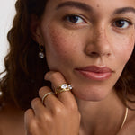 Close-up of a woman wearing gold rings, including a prominent gold ring with a clear gemstone, a delicate gold chain necklace, and a small gold hoop earring, against a clean light background.