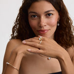 A woman with curly brown hair poses with her hands crossed, showcasing a gold ring with a marquise-cut center stone, a delicate gold bracelet, and a small gold hoop earring with a clear gemstone against a clean white background.