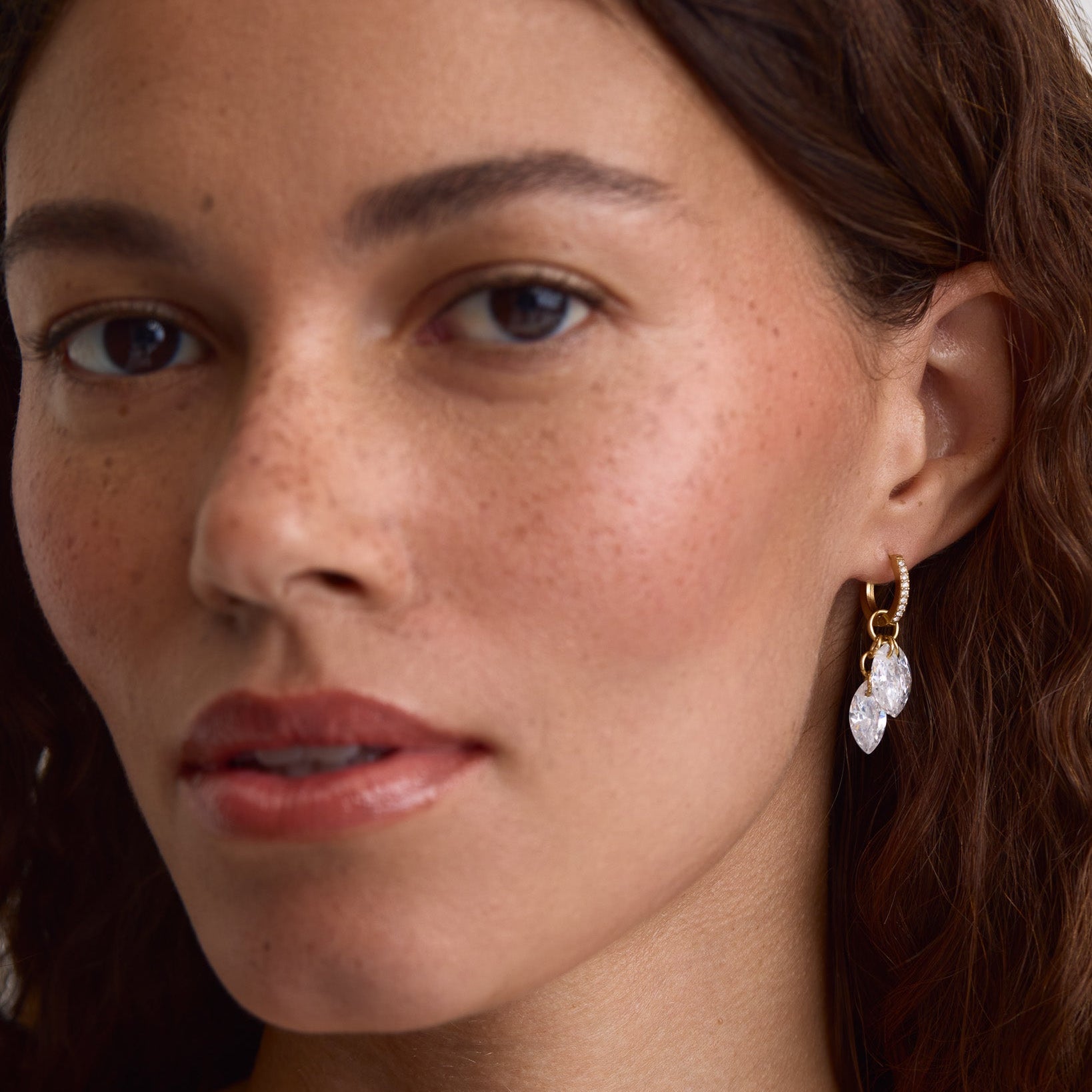 A close-up of a woman wearing a delicate gold hoop earring with a clear faceted drop pendant, shown against her ear and hair.