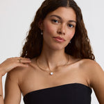 A woman with long, wavy brown hair models a delicate gold chain necklace with a small, round white pearl pendant. She is wearing a black strapless top against a clean, light gray studio background.