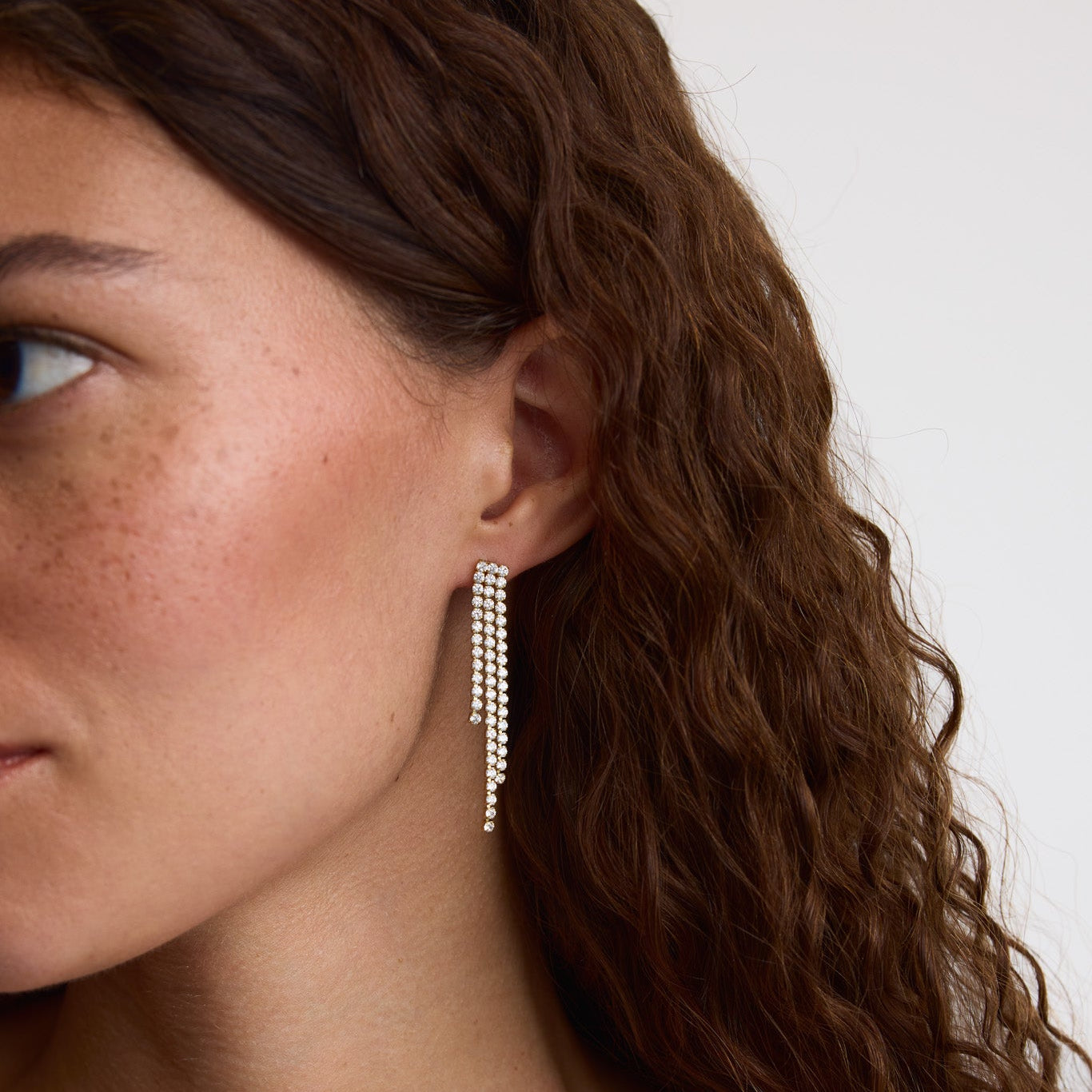 A close-up of a woman with wavy brown hair wearing a long, dangling silver earring with multiple rows of small clear stones against a plain light background.