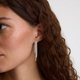 A close-up of a woman with wavy brown hair wearing a long, dangling silver earring with multiple rows of small clear stones against a plain light background.