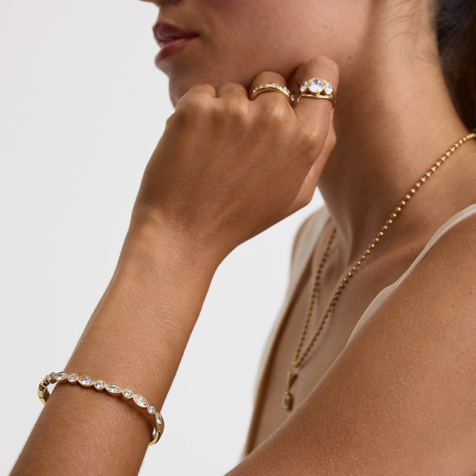 A close-up lifestyle shot of a woman wearing gold jewelry, including a gold bracelet with clear stones on her wrist, a gold ring on her finger, and a gold stud earring, against a clean light gray background.