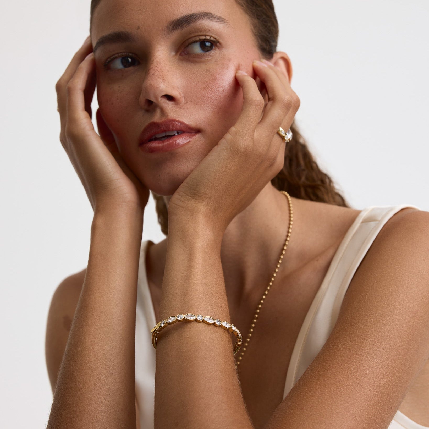 A close-up portrait of a woman with her hands resting on her cheeks, wearing delicate gold jewelry including a gold chain necklace, a gold bracelet, and a gold ring with a clear gemstone, set against a clean white background.