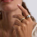A close-up lifestyle shot of a model wearing two gold rings. The primary ring features a large oval-cut clear gemstone in a bezel setting, while the band ring has a channel-set pattern of small clear round gemstones.