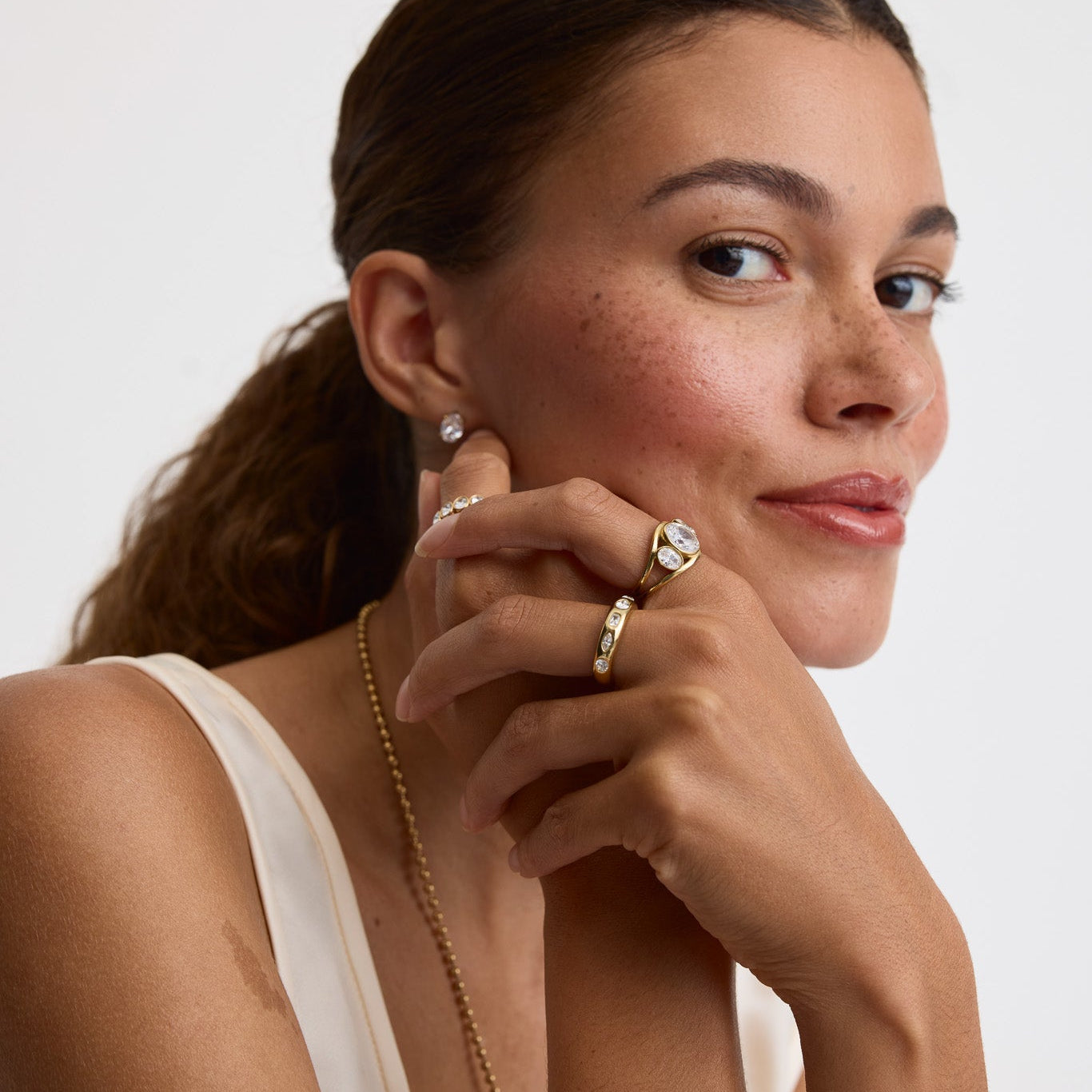 A close-up portrait of a woman with her hair in a ponytail, wearing gold jewelry including a delicate gold chain necklace, a gold ring with a clear stone, and a small gold stud earring, against a plain light gray background.