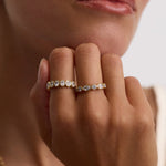 A close-up of a person's hand wearing two delicate gold rings with clear gemstones, set against a soft neutral background.